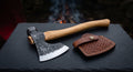 Hand forged bushcraft axe with a curved 19 inch hickory handle and a stamped leather head cover